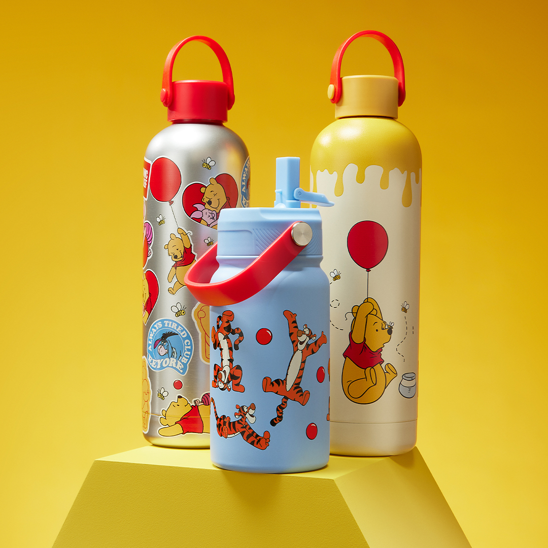 Three Winnie the Pooh–themed water bottles displayed against a bright yellow background. One silver bottle features characters with red balloons, one cream bottle has a honey drip design and Winnie-the-Pooh holding a red balloon, and a smaller blue bottle shows Tigger bouncing among red dots.